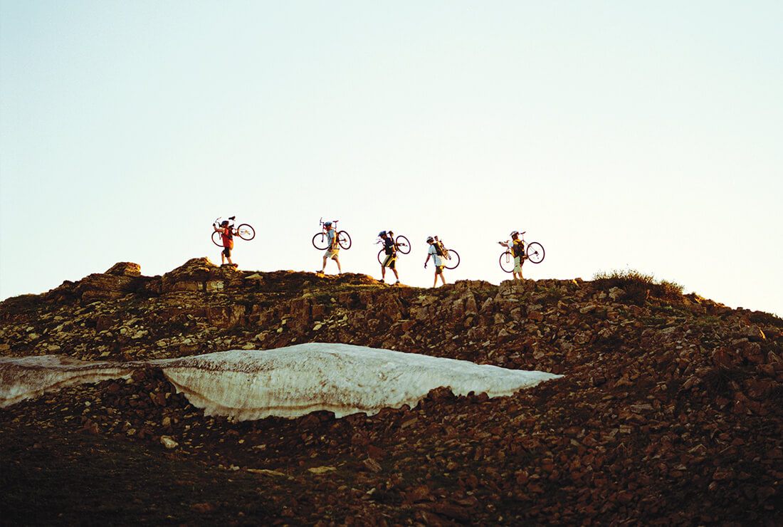 five cyclists carrying their crank brother's bikes and walking along a ridge